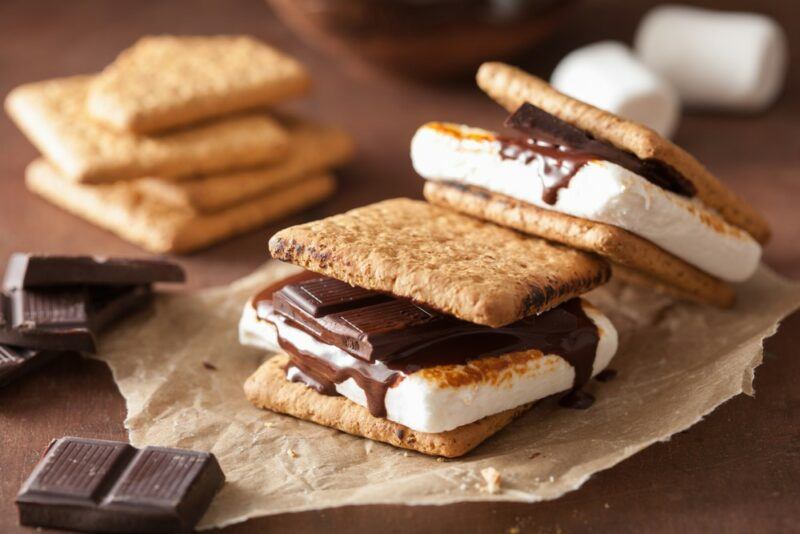 Two decent sized s'mores made using chocolate, marshmallow, and graham crackers, with pieces of chocolate, marshmallows, and more crackers in the background