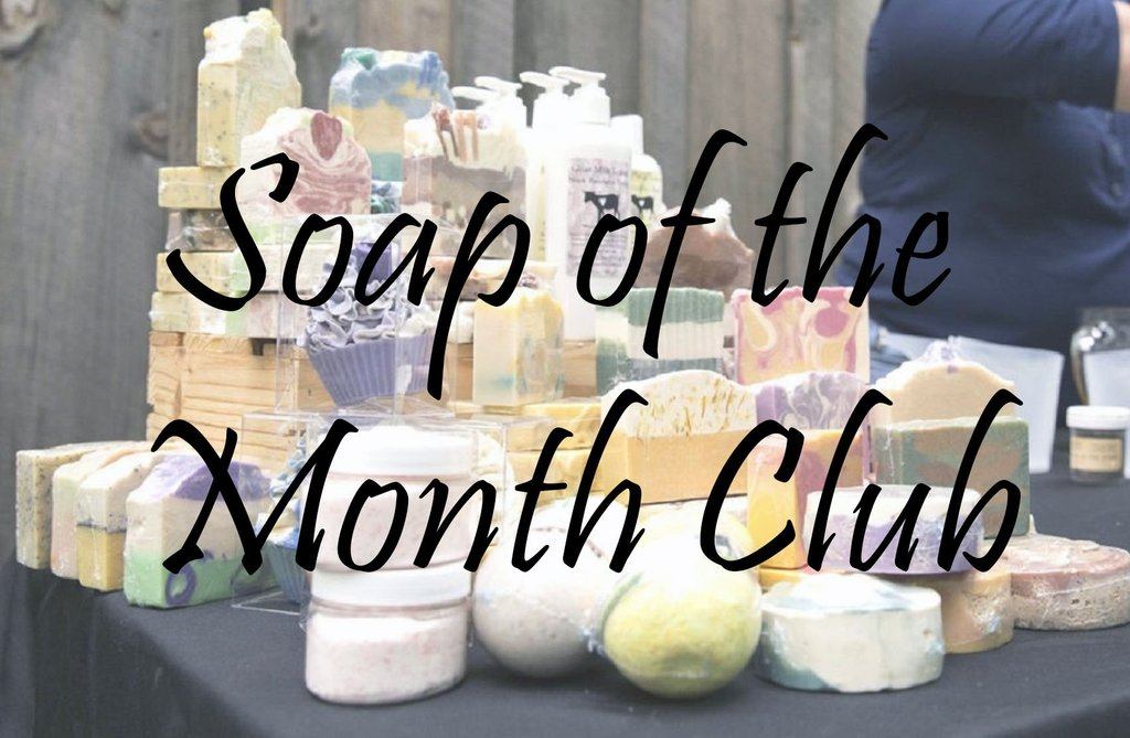 Variety of pastel colored soaps, with lotion bottles, and candles all arranged on a table with black text written across the photo that states, Soap of the Month Club