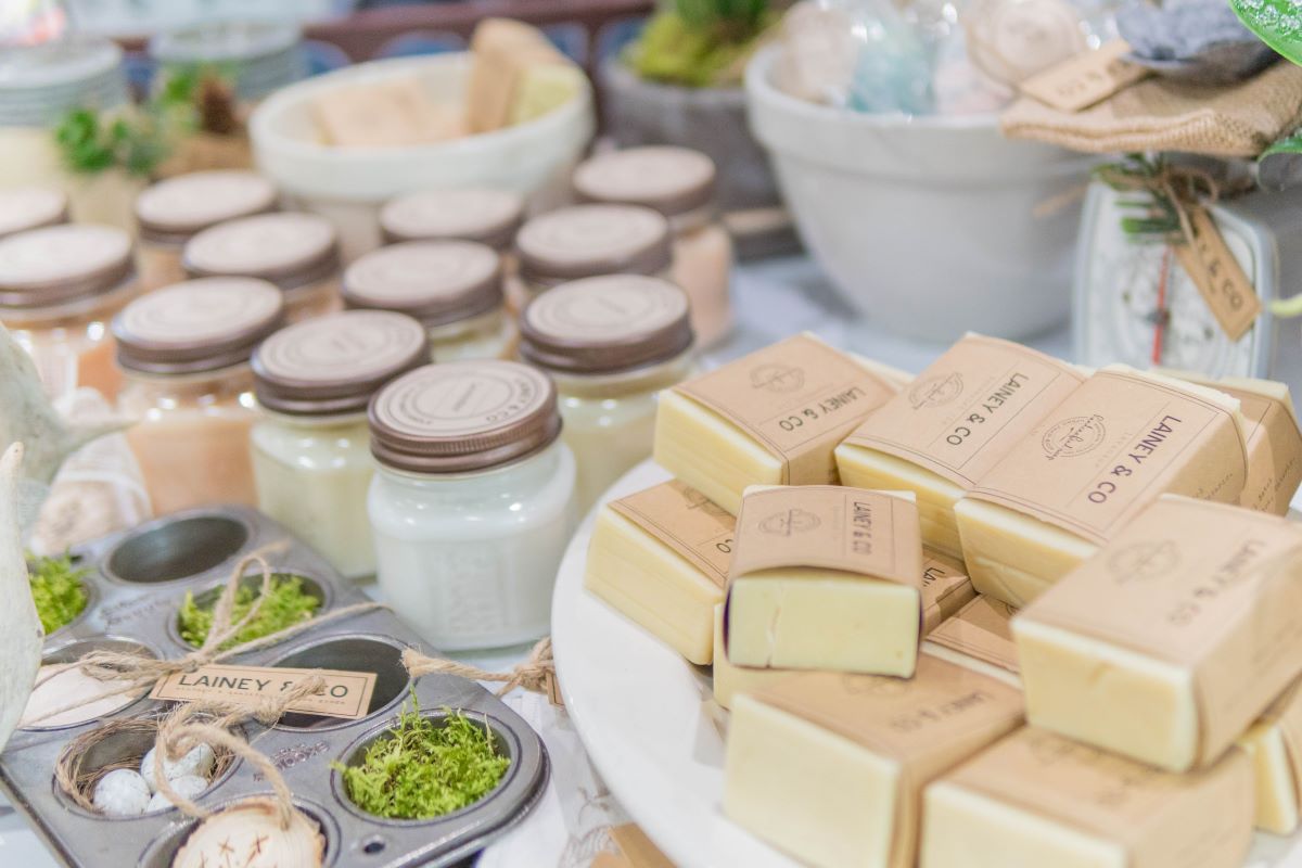 Table at a market with various personal body products, and candles.  Included on the table are artisan soaps.