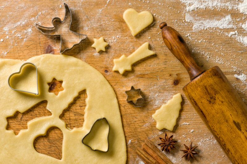 A rolled out piece of cookie dough that's being cut into with different cookie cutters, next to a rolling pin