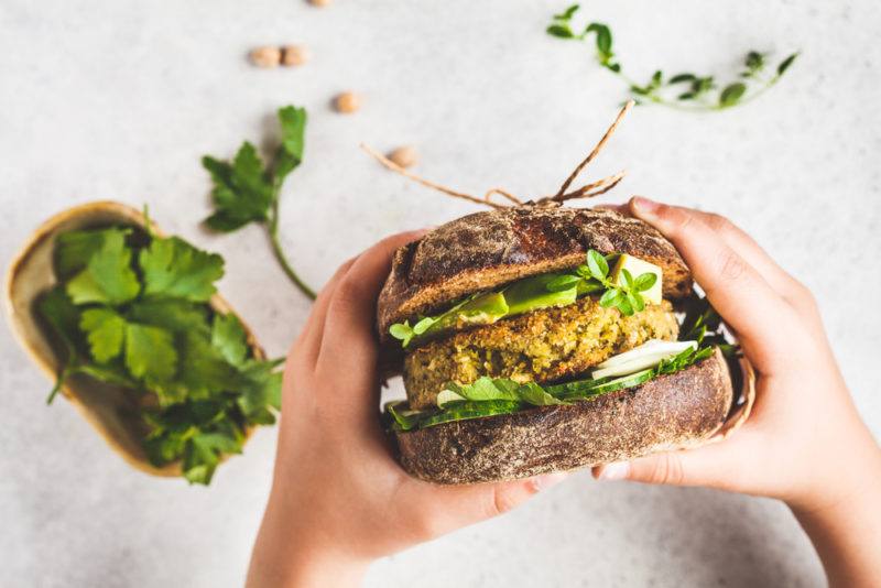 Someone eating a sandwich that includes vegetables and a vegan burger patty, with a white table and some greens in the background