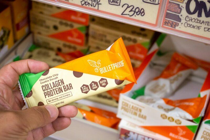 Someone in a grocery store holding a collagen protein bar in their hands, in front of boxes of protein bars