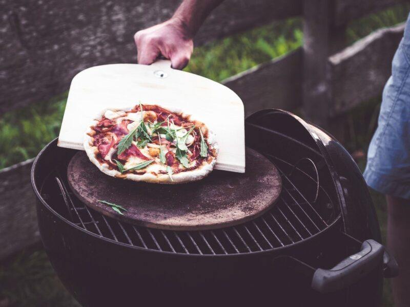 Someone outside using a pizza peel on a pizza stone on a grill. The pizza has all its toppings and looks delicious