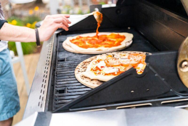 A grill with two pizza stones. One of the stones has a full pizza that's being cooked, while the other has pizza dough that someone is adding sauce too