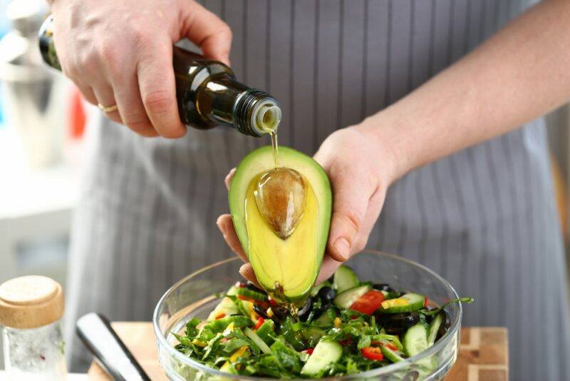 Someone holding an avocado over a bowl of salad and pouring oil onto it