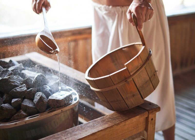 A man wearing a towel scooping water onto coals from a bucket in a sauna