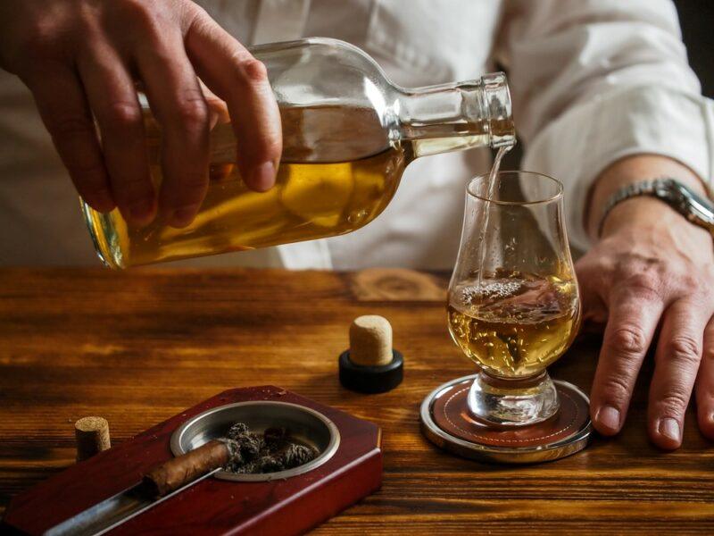 Someone pouring bourbon into a glass next to a cigar in a cigar tray