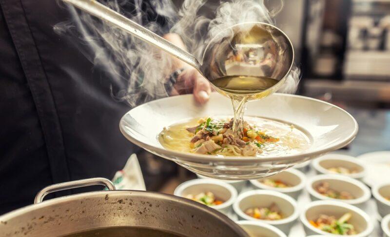 A chef spooning chicken broth with a ladle into bowls