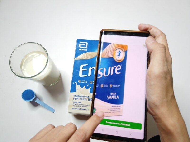 Someone taking a photo of Ensure using a smartphone, next to a glass and a scoop for the protein powder