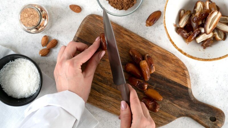 A woman in the kitchen, cutting a date on a wooden board, which will be used to make bliss balls