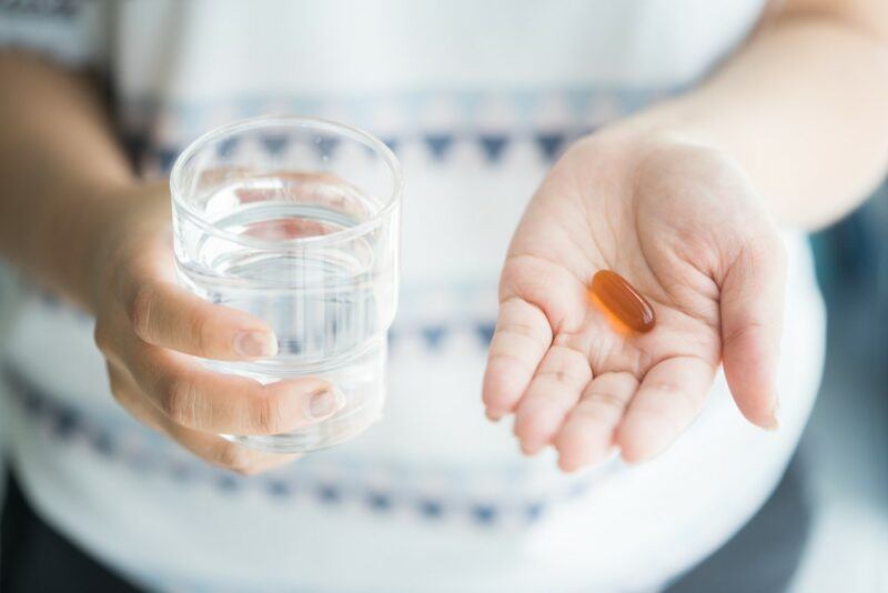 A woman in a patterned shirt, holding a glass of water in one hand and a fish oil supplement in the other