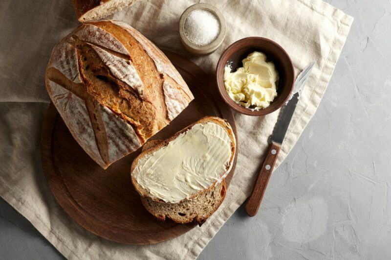 A loaf of sourdough bread, with one slice of bread buttered, next to a small jar butter