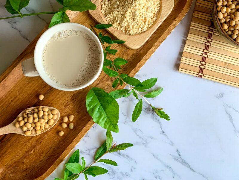 A wooden tray with a mug of soy milk, soy beans on a spoon, soy powder, and leaves