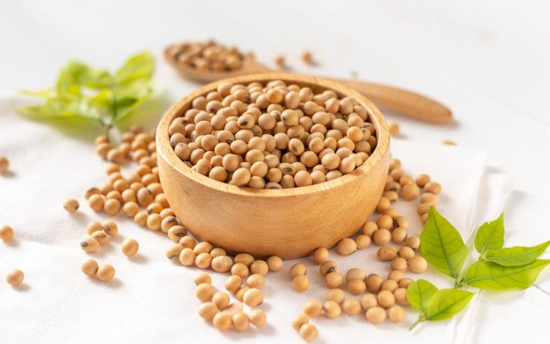 A wooden bowl containing soybeans, with soybeans scattered on a white table