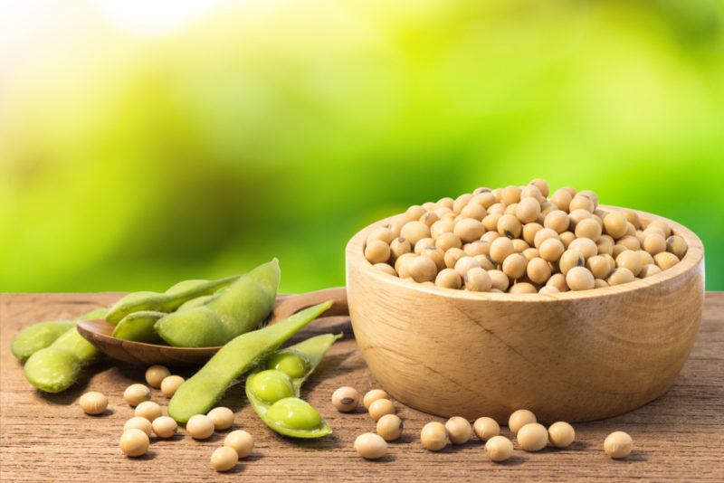 A wooden bowl of shelled soybeans, with whole soybeans and some more shelled ones next to it on a table. The background looks like the photo is taken outdoors.