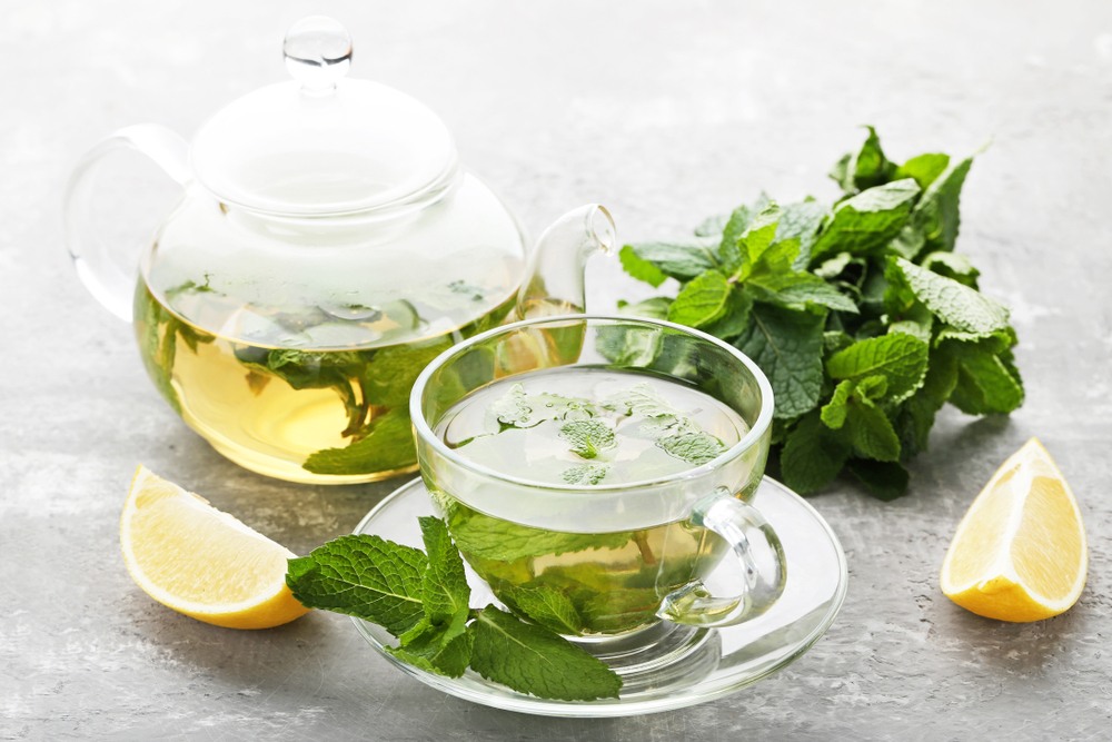 A glass mug of spearmint tea, next to a glass pot of the tea, some spearmint leaves and lemon wedges