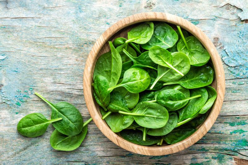 A wooden bowl filled with raw spinach leaves rests on a rustic wooden surface near three loose spinach leaves.