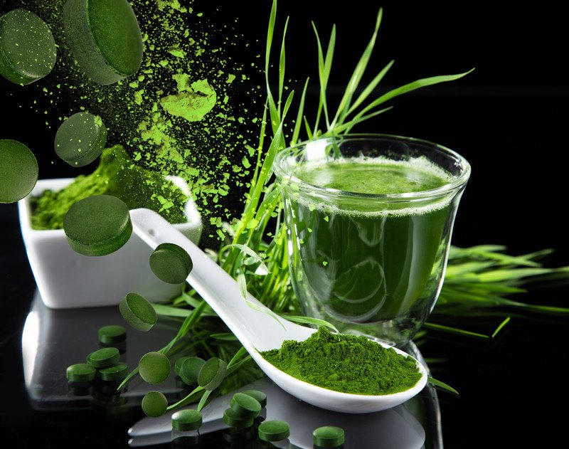 This photo shows powdered spirulina, pills of spirulina, a glass of spirulina in water, and a white spoon with spirulina powder on a dark background.