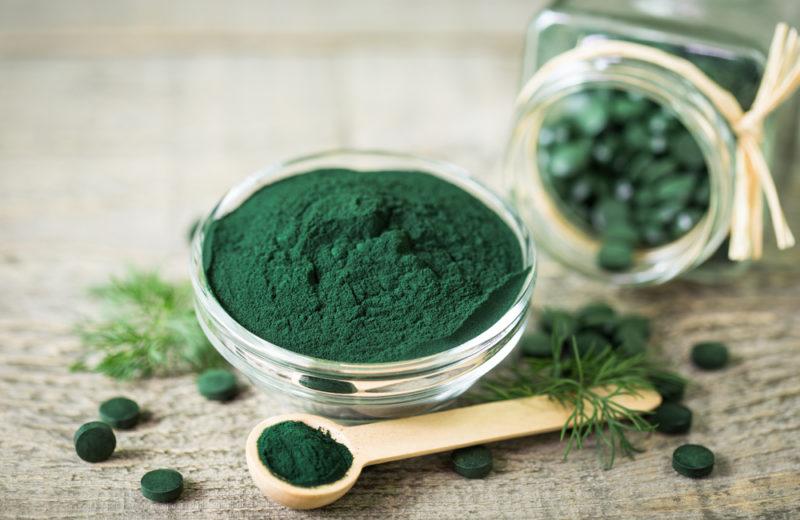 A glass bowl of spirulina, with a spoon and a glass jar containing spirulina tablets