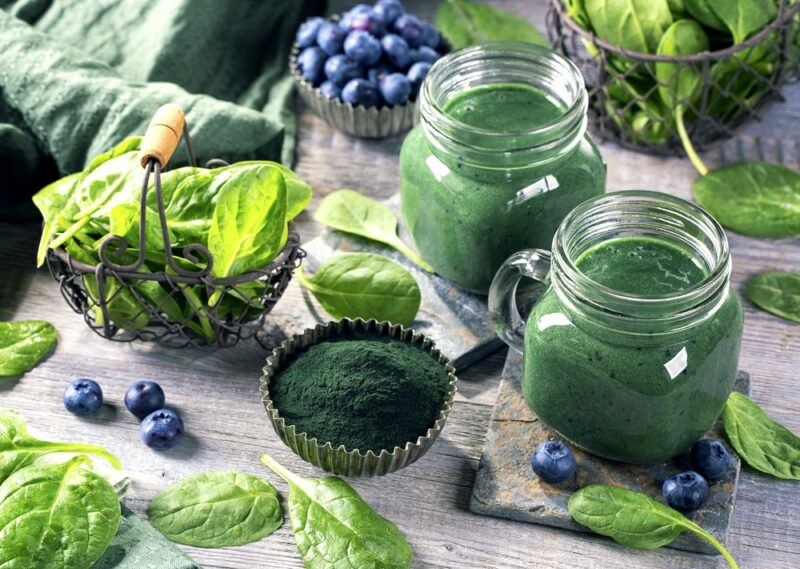 Two spirulina smoothies, with various ingredients scattered across the table, including spirulina powder, blueberries, and spinach