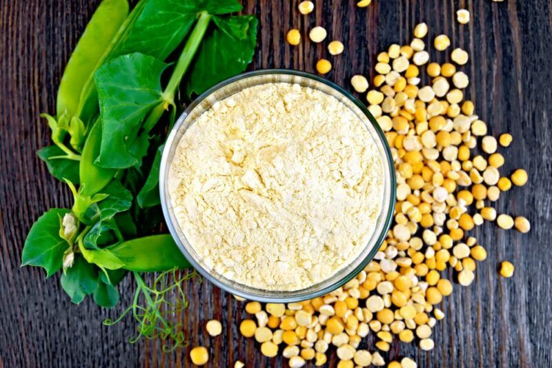 A glass jar of pea protein powder next to split yellow peas and some leaves