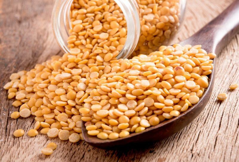 A wooden table with a jar of split peas spilling onto the table, next to a large spoon of the peas