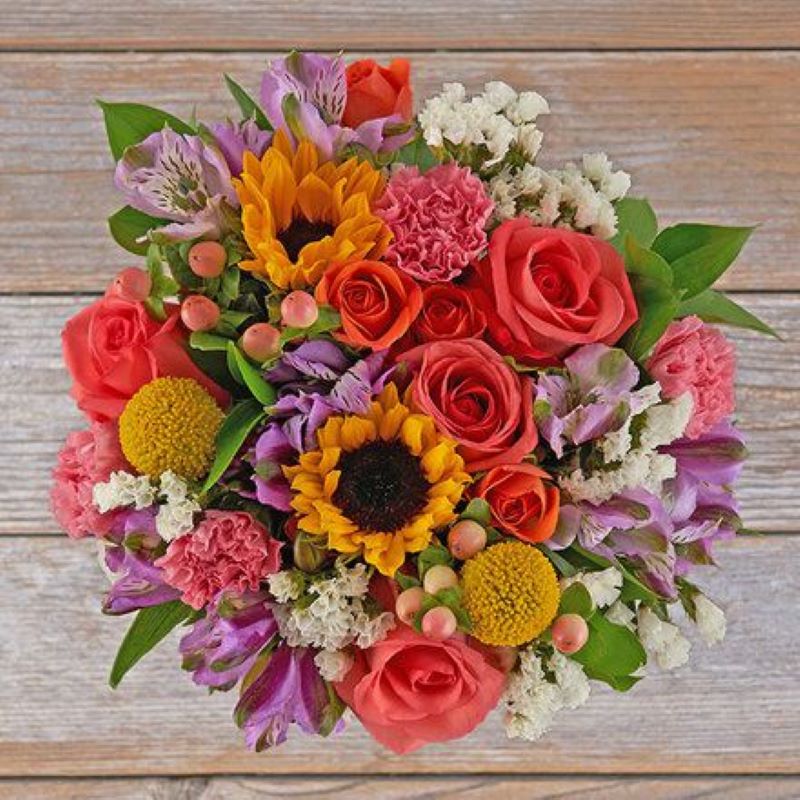 Large Summer bouquet with sun flowers, coral roses, and other various colored flowers and berries and greens sitting on a wood table