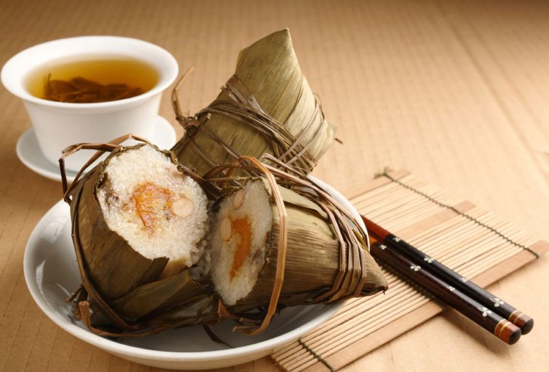 A steamed rice dumpling that has been wrapped in bamboo leaves and another cut in half, in front of a mug of tea