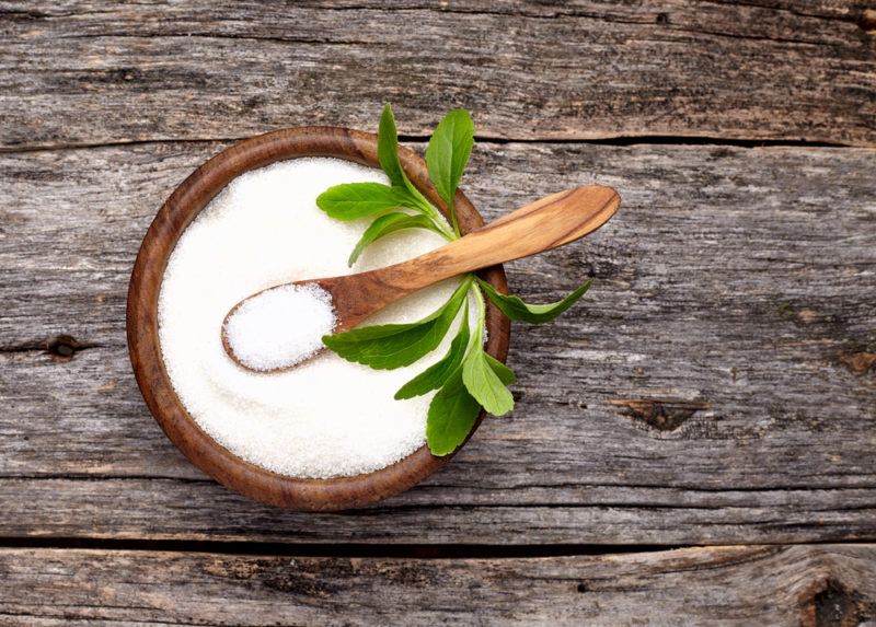 A brown bowl of stevia with a spoon and stevia leaves on a wooden table