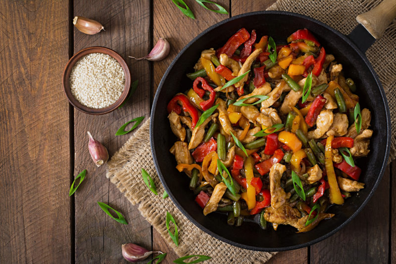 A black frypan on a wooden table that contains a fresh stirfry with peppers and other ingredients