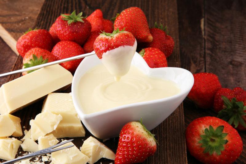 A heart shaped white container of white chocolate, with white chocolate and strawberries around it. One strawberry is being dipped into the chocolate.