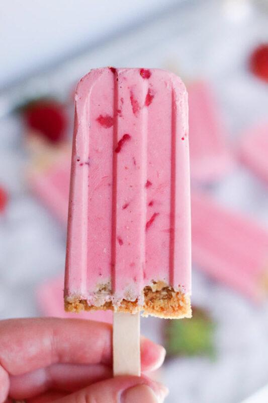 Someone holding a strawberry cheesecake popsicle with graham cracker crust