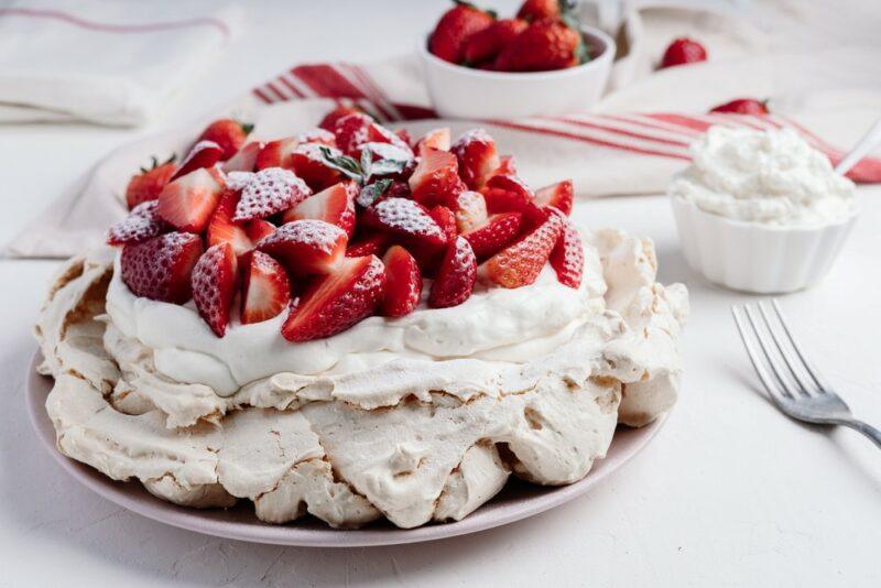 A large pavlova loaded with strawberries and cream, with a small bowl of strawberries and one of cream in the background