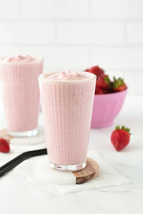 Two glasses containing a strawberry smoothie made with almond milk, with a pink bowl of strawberries in the background