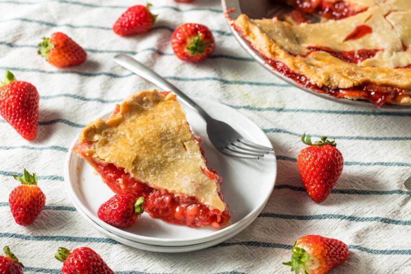 A piece of strawberry pie on a white plate, with strawberries scattered around it and the rest of the pie in the background