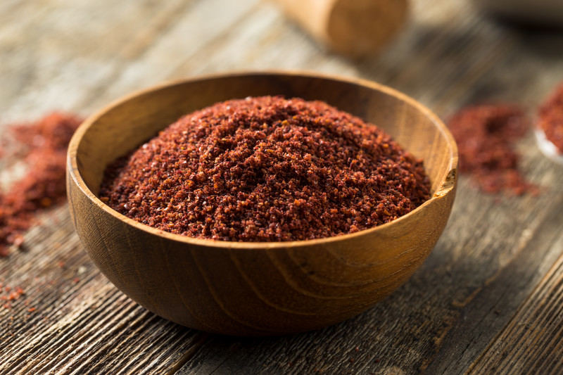 Wooden bowl with a chipped rim full of coarsely ground sumac, laid out on a wooden table with loose sumac powder at the back.