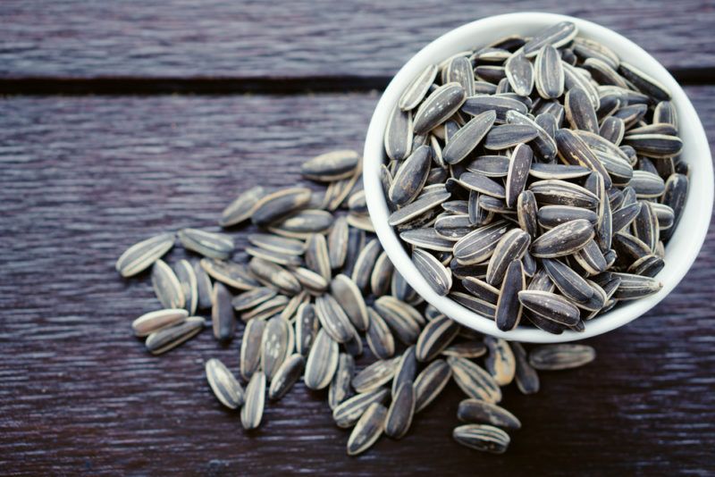 A white bowl filled with sunflower seeds, with more seeds below it on a dark wooden table