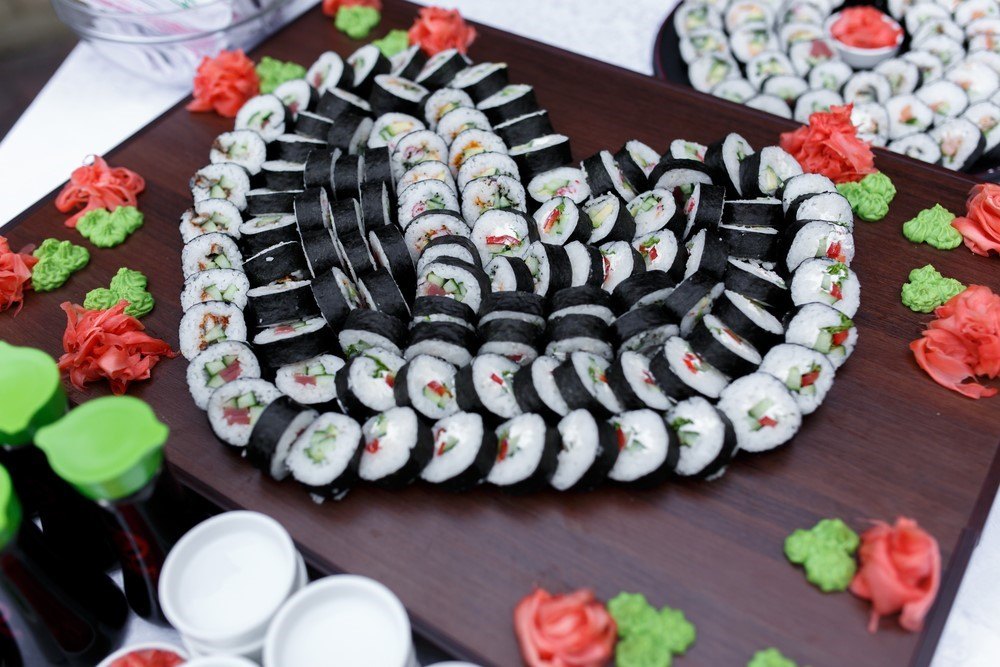 A large wooden board with sushi in the shape of a heart, with ginger and wasabi flowers