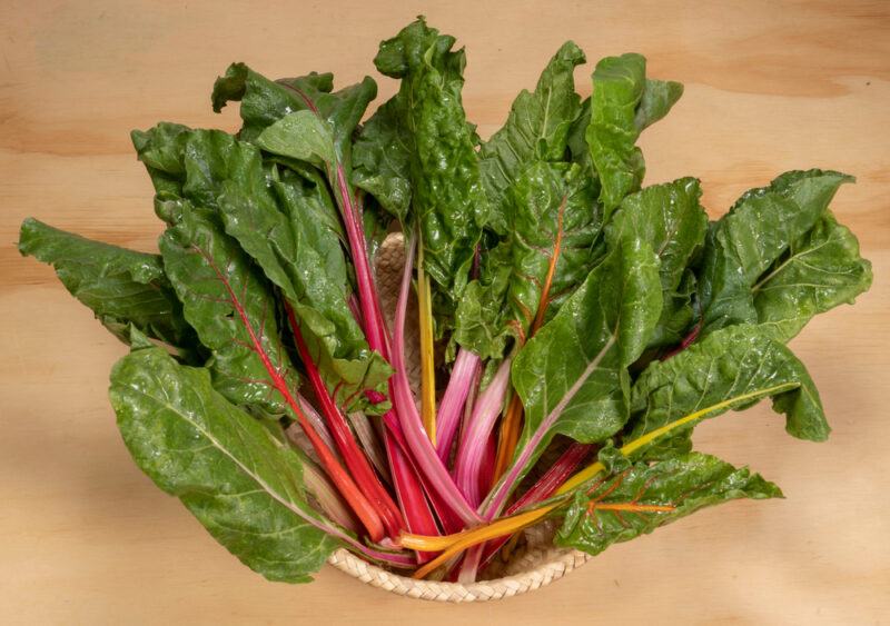 on a light brown wooden surface is a weaved bowl full of Swiss chard