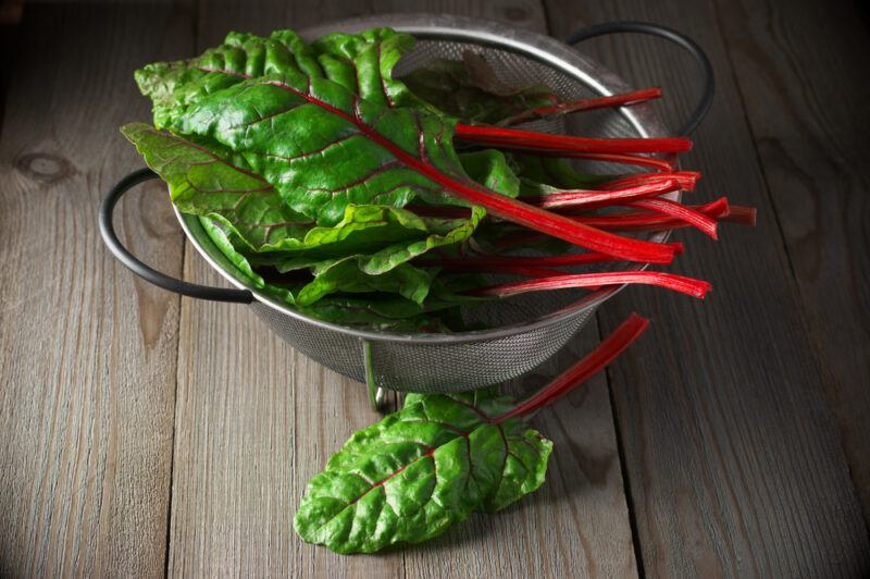 on a wooden surface is a metal colander with two handles full of Swiss chard, with a piece of Swiss chard in front of it