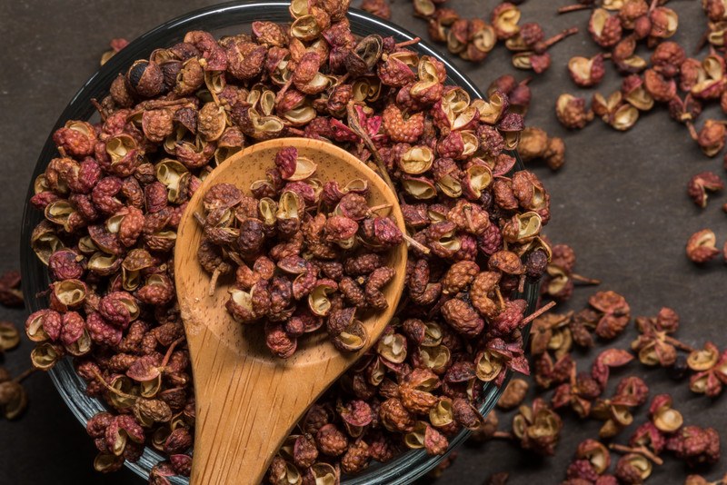 top view image of Szechuan peppers on a black plate with wooden spoon, resting on a dark-colored surface