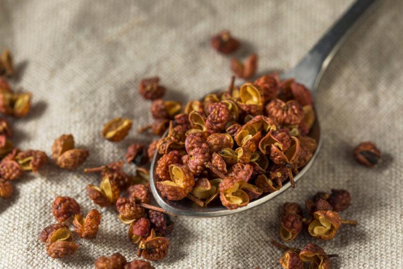 on a tan colored kitchen towel is a closeup image of a silver spoon full of Szechuan peppercorns with some of it spilled around