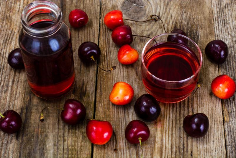 A collection of tart cherries on a table, along with a bottle and a cup of tart cherry juice