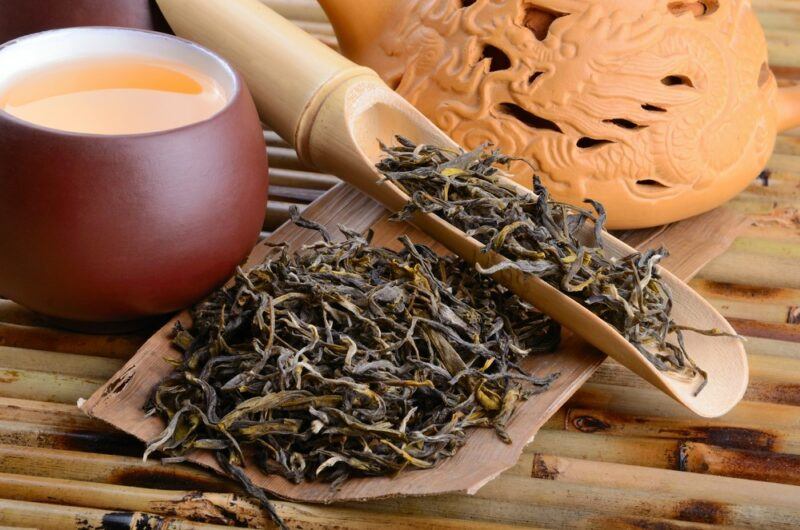 A wooden board with tea leaves and another scoop of leaves, next to a mug of tea