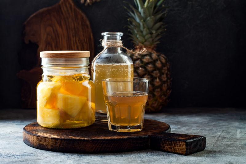 A glass, a bottle and a jar containing Tepache, next to a pineapple