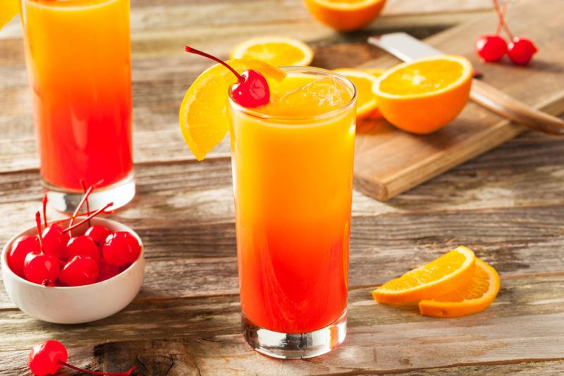 Two tequila sunrise cocktails on a table with glace cherries and oranges