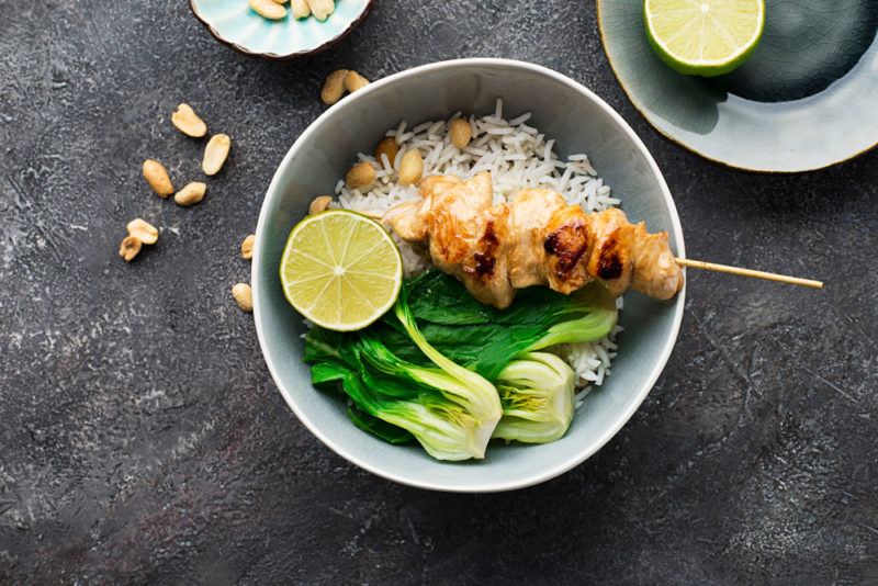 A white bowl with a chicken skewer, rice and bok choy