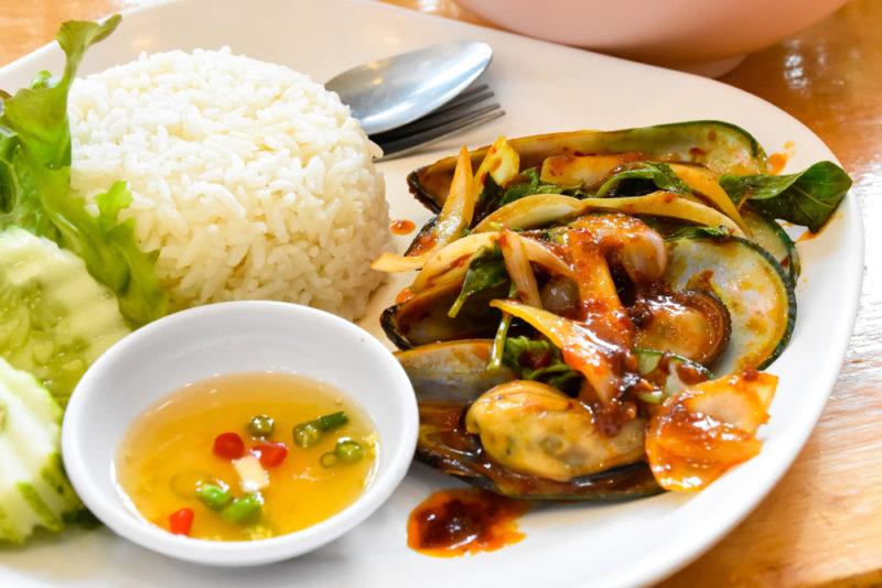 A white plate with Thai style mussels, next to rice, a spoon, veggies and dipping sauce