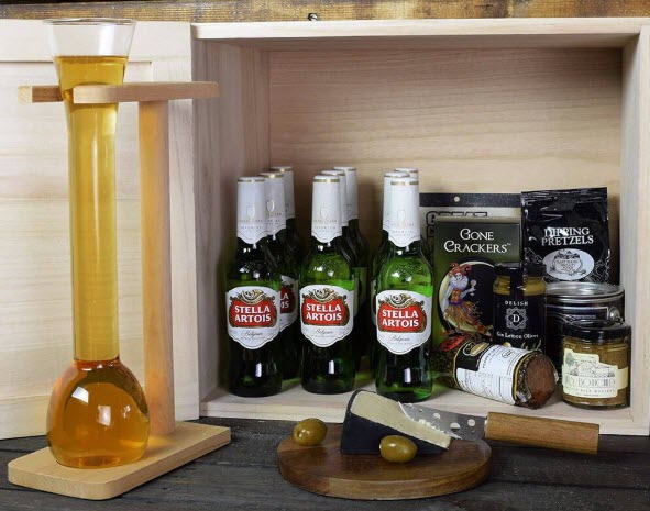 An open crate containing 9 bottles of Stella Artois and plenty of snacks. There is a full half yard beer glass next to it, along with a cutting board, cheese and olives. 