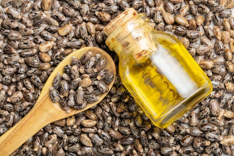 A large pile of the seeds used to make castor oil, with a spoon of the seeds on top and a small bottle of castor oil